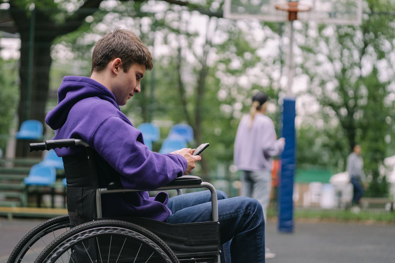 A young man in a wheelchair using a smartphone in an outdoor park setting.